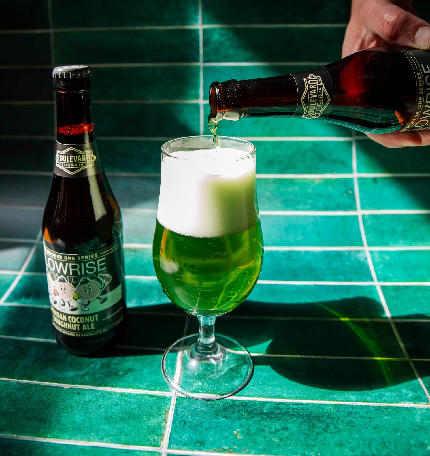 A hand pours a bottle of golden bubbly beer into a glass with frothy white head, set against green tiled background. Another beer bottle stands nearby, labeled with a playful design.